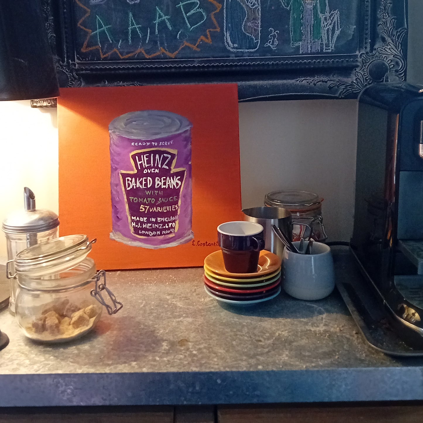Kitchen counter with jars, a cup, and a painting of a purple can of Heinz baked beans on an orange board.